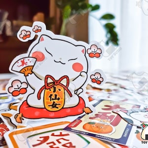 May include: A white cartoon cat with red cheeks holding a red and gold fan. The cat is wearing a red and gold scarf and a red and gold pouch with the text "小仙女" in black. The cat is sitting on a red cushion with a white border. The background is a table with a variety of colorful stickers.