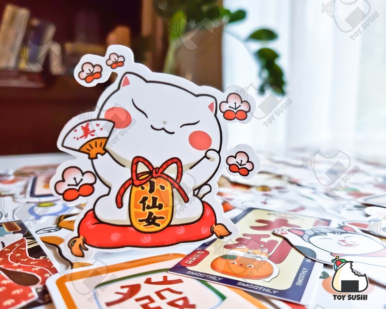 May include: A white cartoon cat with red cheeks holding a red and gold fan. The cat is wearing a red and gold scarf and a red and gold pouch with the text "小仙女" in black. The cat is sitting on a red cushion with a white border. The background is a table with a variety of colorful stickers.