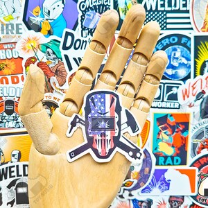 May include: A sticker featuring a skull wearing a welding mask, with an American flag design. The sticker is held by a wooden hand, surrounded by other welder-themed stickers. The design includes crossed tools and patriotic colors.