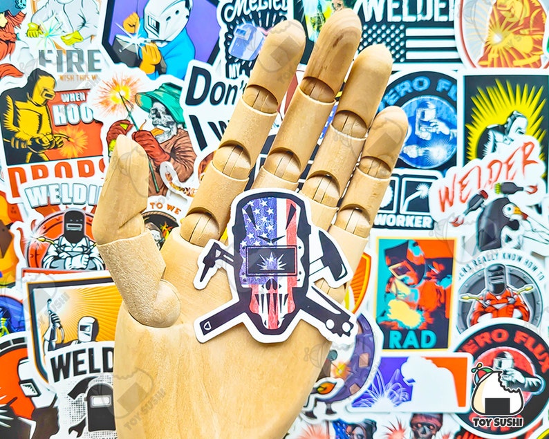 May include: A sticker featuring a skull wearing a welding mask, with an American flag design. The sticker is held by a wooden hand, surrounded by other welder-themed stickers. The design includes crossed tools and patriotic colors.