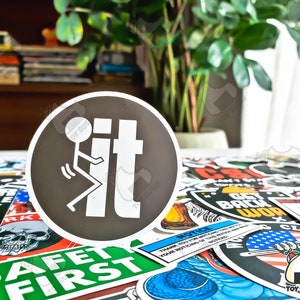 May include: A black and white sticker with a stick figure flipping the bird and the word "it" in white letters. The sticker is on a table with other stickers.