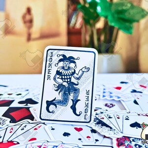 May include: A playing card with a joker design. The joker is depicted as a jester with a black and white harlequin costume. The card is white with black text that reads "JOKER" on the left side and "JOKER" vertically on the right side.