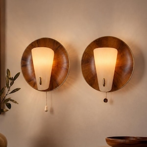 May include: Two wall-mounted sconces featuring circular wooden backplates with a rich grain pattern. Each light has a frosted, cone-shaped glass shade and a small brass accent. A pull cord hangs from the bottom of each fixture for operation.