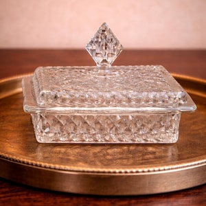 Könnte beinhalten: Eine klare, quadratische Glas-Schmuckschatulle mit einem rautenförmigen Muster. Der Deckel hat einen rautenförmigen Griff. Die Box wird auf einem goldfarbenen Tablett präsentiert und zeigt ihr elegantes Design.