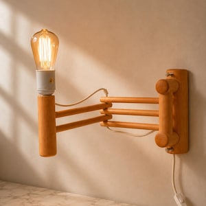 Può includere: Una lampada da parete con braccio a fisarmonica in legno chiaro e base verticale in legno. L'apparecchio presenta un portalampada bianco con una lampadina a filamento a vista che emette una luce calda e un cavo bianco con interruttore.