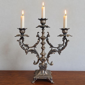 May include: A bronze-toned metal candelabra featuring three arms with ornate, scrolling leaf and vine details. The central stem supports three lit, off-white taper candles held in petal-shaped cups. The base is square with decorative feet.