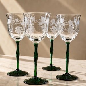 Könnte beinhalten: Ein Set aus vier Weingläsern aus klarem Glas mit geätzten weißen Trauben- und Weinrankenmustern an den Kuppen. Jedes Glas hat einen schlanken, massiven dunkelgrünen Stiel und einen passenden dunkelgrünen runden Fuß als Kontrast zum klaren Glas.