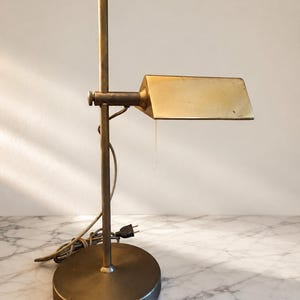 Könnte beinhalten: Eine Schreibtischlampe aus Messing mit rundem Fuß, vertikalem Stab und verstellbarem horizontalem Arm mit rechteckigem Schirm. Die Lampe verfügt über einen Zugschalter sowie ein Stromkabel mit Stecker und hat ein metallisches Finish.