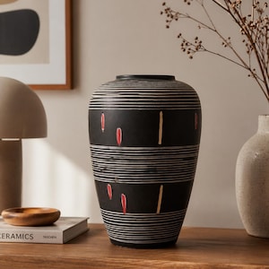 Puede incluir: Un jarrón de cerámica negro con rayas blancas horizontales y detalles verticales rojos y dorados. El jarrón tiene forma cilíndrica y se exhibe sobre una superficie de madera, mostrando un diseño moderno y minimalista.