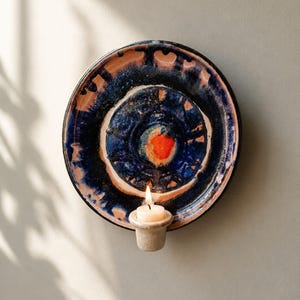 Könnte beinhalten: Eine kreisförmige Wandleuchte aus Keramik mit einer tiefblauen und schwarzen Glasur und einem markanten orangefarbenen Zentrum. Das Stück verfügt über einen integrierten beigen Kerzenhalter am unteren Rand mit einem brennenden Teelicht.
