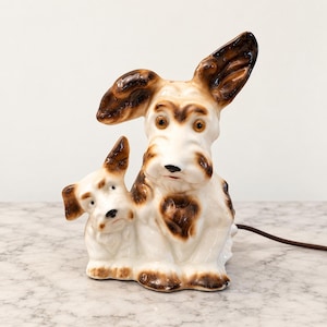 May include: A ceramic table lamp shaped like two dogs sitting side by side. The dogs are white with brown patches on their ears, faces, and bodies. The larger dog has one ear standing upright and one folded down. A brown electrical cord extends from the back.