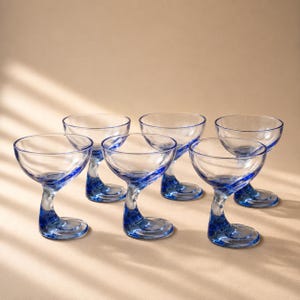 Könnte beinhalten: Ein Set aus sechs klaren Glasschalen, die jeweils einen markanten, geschwungenen Fuß aus kobaltblauem Glas aufweisen. Die Schalen sind breit und flach, für das Servieren von Cocktails oder Desserts konzipiert, mit einer glatten, transparenten Oberfläche.