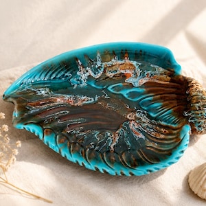 Könnte beinhalten: Dekorative Keramikschale in Muschelform mit türkisfarbener und brauner Glasur. Die Schale hat eine strukturierte Oberfläche und ein detailliertes Design. Neben der Schale liegt eine kleine Muschel.