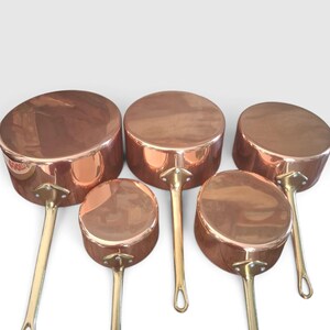 Op de afbeelding: Een set van vijf gepolijste koperen steelpannen van verschillende groottes, elk met een ronde vorm en een lange, slanke messing steel bevestigd met een driehoekige metalen plaat met drie klinknagels. De pannen zijn in een groep geplaatst.