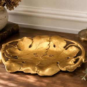 Könnte beinhalten: Eine goldfarbene dekorative Schale in Form von zwei sich überlappenden Blättern. Die Schale hat aufwendige Aderdetails und ein geschwungenes Griffdesign. Sie steht auf einer Holzoberfläche, mit einem Kerzenhalter und einer Vase im Hintergrund.