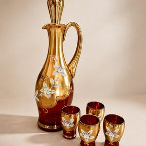 Könnte beinhalten: Ein Vintage-Dekanter-Set in Gold und Rubinrot. Der Dekanter hat einen Griff und einen Verschluss mit floralen Akzenten. Vier passende Schnapsgläser sind ebenfalls enthalten, jedes mit einem goldenen Rand und floralem Design.