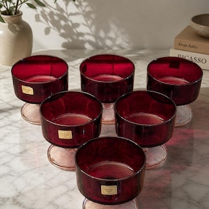 Könnte beinhalten: Sechs rubinrote Dessertschalen aus Glas mit Stiel und rundem Fuß. Die Schalen haben eine zylindrische Form und sind kreisförmig angeordnet. Jede Schale hat ein kleines goldenes Etikett. Die Schalen stehen auf einer Marmoroberfläche.