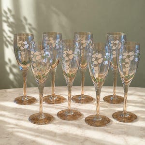 Op de afbeelding: Een set van acht hoge champagneglazen met een lichte amberkleurige tint. Elk glas is voorzien van een delicaat handgeschilderd ontwerp van witte bloemen met groene bladeren en stengels rondom de kelk. De stelen en ronde voeten zijn helder.