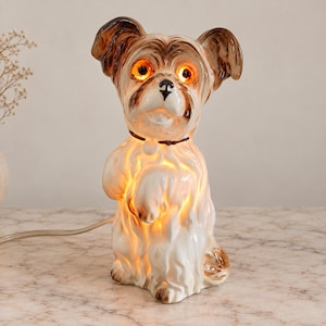 May include: A ceramic dog-shaped lamp with a glossy white finish and brown accents on the ears and face. The lamp features glowing orange eyes and a light source inside the body that illuminates the white fur texture. A thin brown collar is visible.