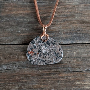 May include: A brown leather cord necklace with a copper wire wrapped pendant featuring a grey, black, and red speckled stone.