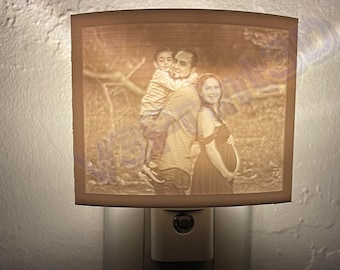 May include: A white night light with a photo of a family of three. The photo shows a man holding a young boy on his shoulders, while a woman stands next to them. The photo is printed on the front of the night light.