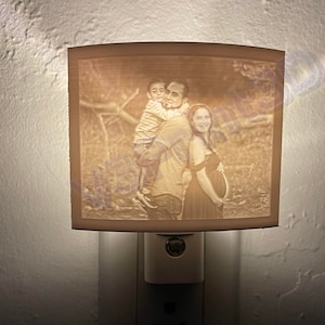May include: A white night light with a photo of a family of three. The photo shows a man holding a young boy on his shoulders, while a woman stands next to them. The photo is printed on the front of the night light.