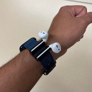 May include: A navy blue watch band with a black clasp holds a pair of white wireless earbuds. The earbuds are attached to a black and silver holder. The band is secured around a wrist.