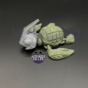 Könnte beinhalten: Eine 3D-gedruckte, olivgrüne Schildkrötenfigur mit detailliertem Panzer und beweglichen Gliedmaßen. Die Schildkröte hat einen silberfarbenen Kopf und einen kleinen, runden "AUTHORIZED SELLER" Aufkleber.