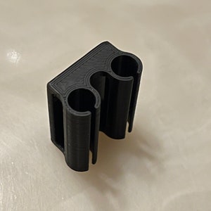May include: A black 3D-printed clip with two circular openings and a rectangular base. The clip is designed to hold two cylindrical objects. The material appears to be a matte plastic.