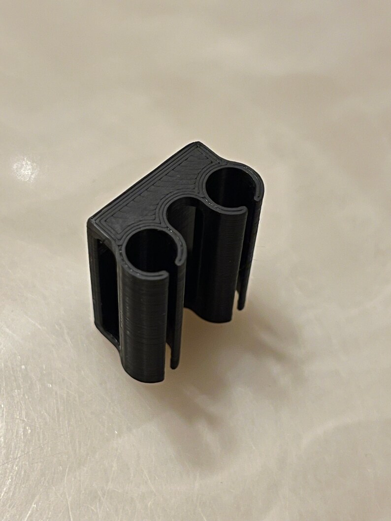 May include: A black 3D-printed clip with two circular openings and a rectangular base. The clip is designed to hold two cylindrical objects. The material appears to be a matte plastic.