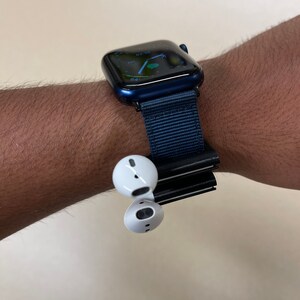 May include: A navy blue smartwatch with a matching fabric band is worn on a wrist. White wireless earbuds are attached to the watch band. The watch face displays a digital interface.