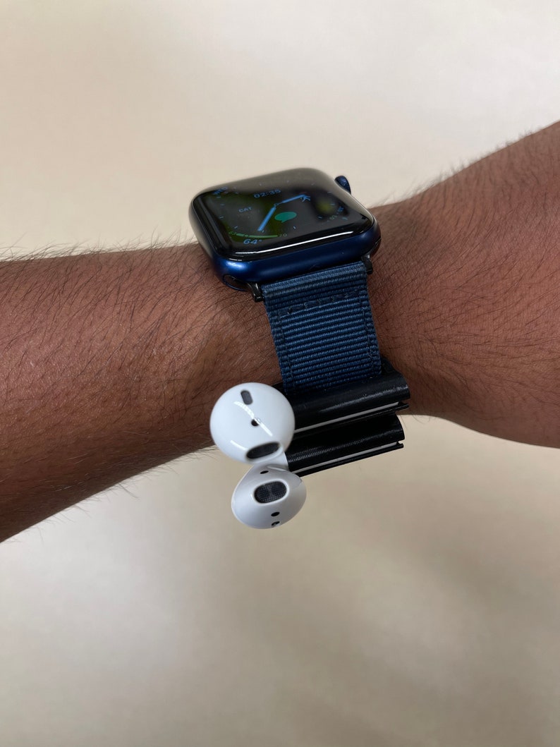 May include: A navy blue smartwatch with a matching fabric band is worn on a wrist. White wireless earbuds are attached to the watch band. The watch face displays a digital interface.
