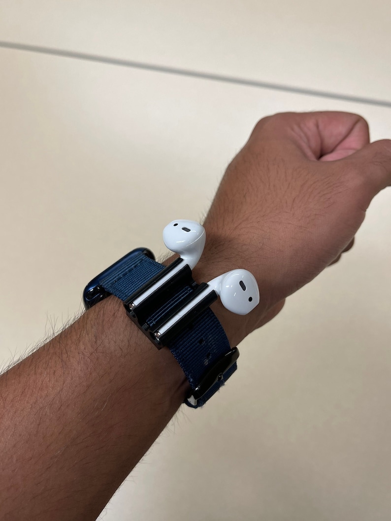 May include: A navy blue watch band with a black clasp holds a pair of white wireless earbuds. The earbuds are attached to a black and silver holder. The band is secured around a wrist.