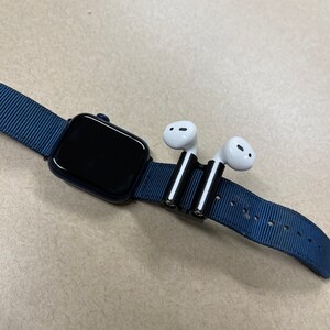 May include: A navy blue Apple Watch with a black screen and a matching blue nylon band. A black and silver holder secures a pair of white wireless earbuds to the watch band.