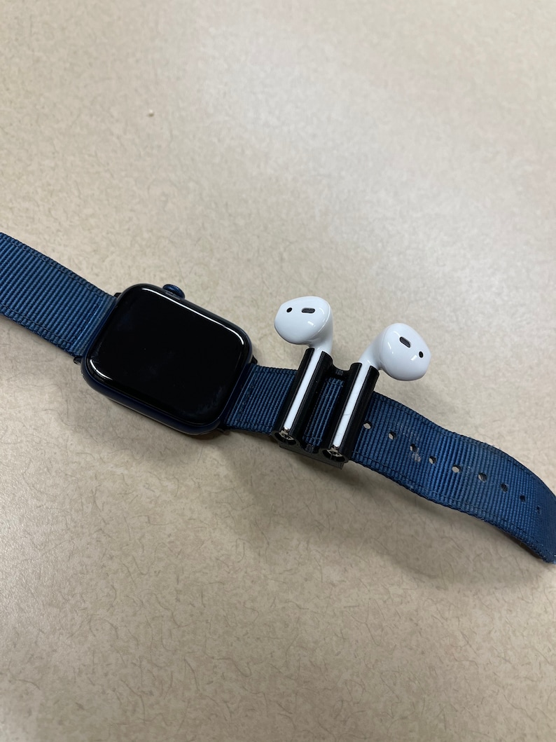 May include: A navy blue Apple Watch with a black screen and a matching blue nylon band. A black and silver holder secures a pair of white wireless earbuds to the watch band.