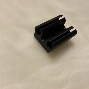 May include: A small, black, rectangular object with two holes and three slots. The object appears to be made of a solid material and has a matte finish. The item is likely a component or part of a larger product.