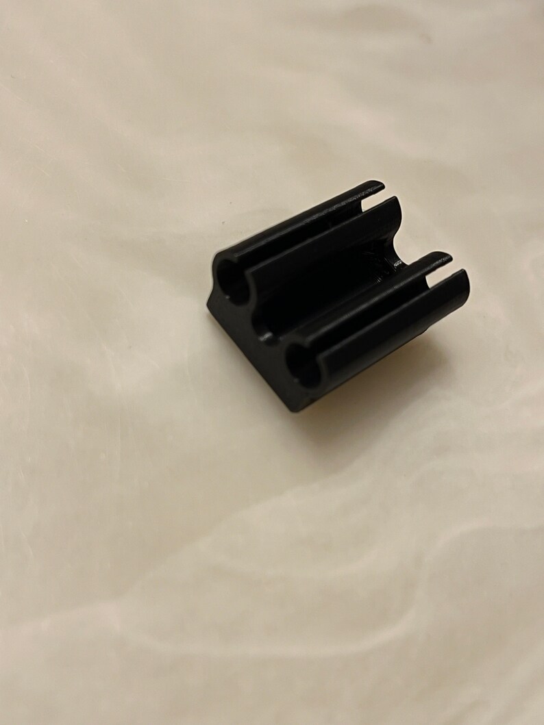 May include: A small, black, rectangular object with two holes and three slots. The object appears to be made of a solid material and has a matte finish. The item is likely a component or part of a larger product.