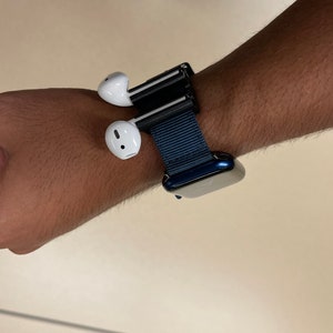 May include: A wrist-worn accessory featuring a dark blue watch band and a black holder for white wireless earbuds. The earbuds are positioned on the side of the wrist, offering a unique way to carry them.