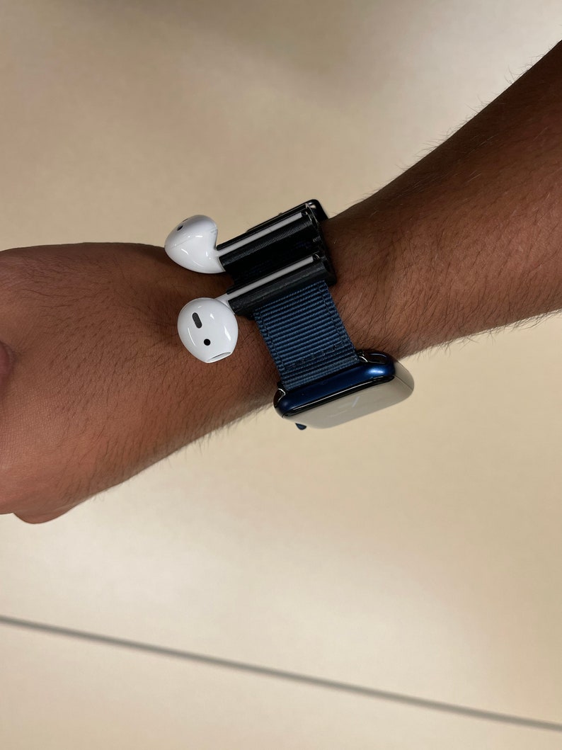 May include: A wrist-worn accessory featuring a dark blue watch band and a black holder for white wireless earbuds. The earbuds are positioned on the side of the wrist, offering a unique way to carry them.