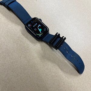 May include: A navy blue smartwatch with a matching nylon band and black buckle. The watch face displays the time, date, and activity data. A black accessory is attached to the band.