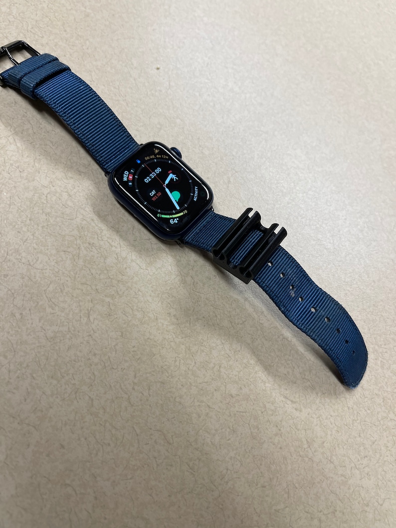 May include: A navy blue smartwatch with a matching nylon band and black buckle. The watch face displays the time, date, and activity data. A black accessory is attached to the band.