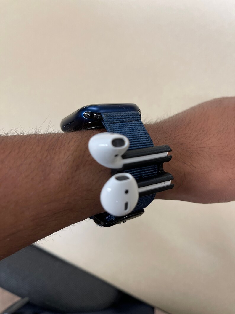 May include: A navy blue Apple Watch with a matching fabric band. Two white wireless earbuds are attached to the band. The watch and earbuds are on a wrist.