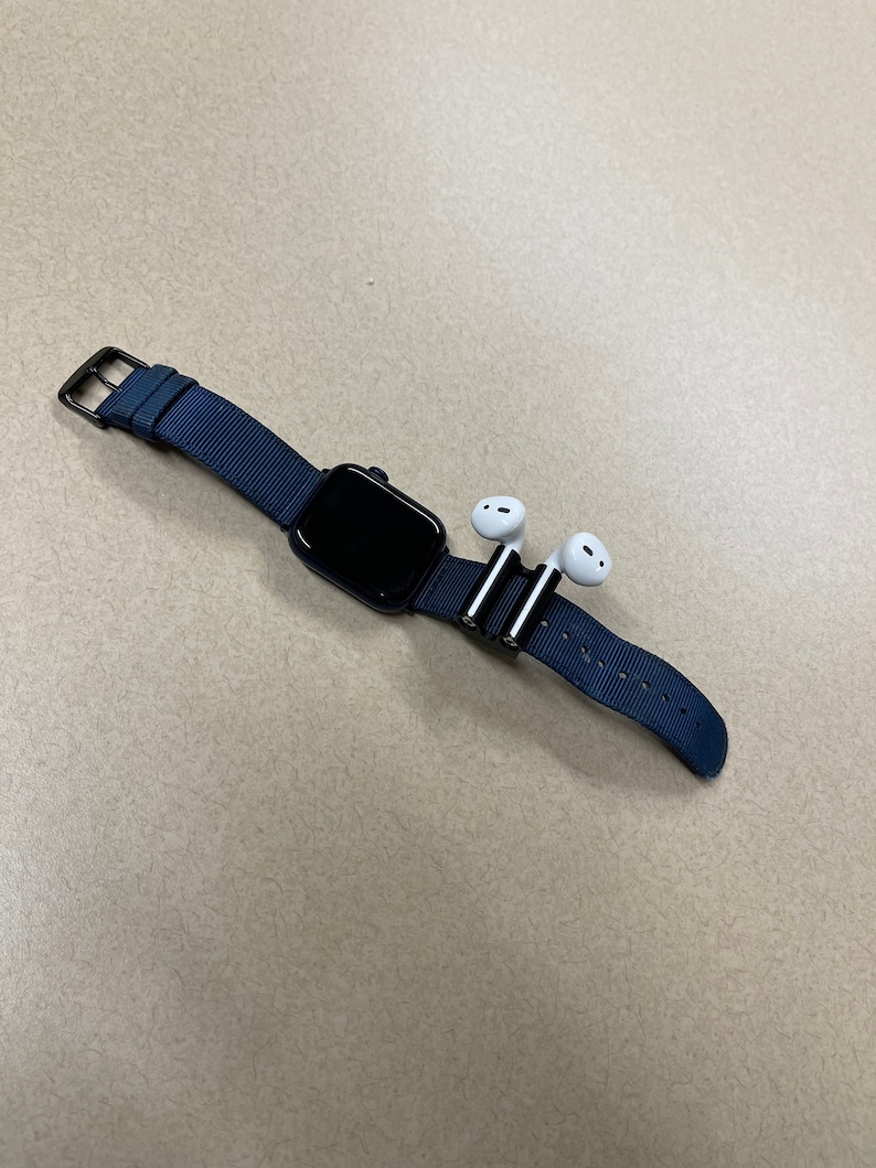 May include: A black smartwatch with a navy blue fabric band and a black buckle. Two white wireless earbuds are attached to the band with silver clips. The watch is displayed on a neutral-toned surface.