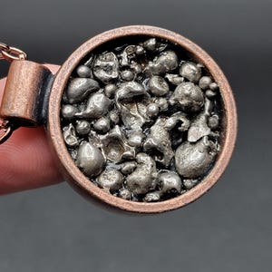 May include: A copper pendant necklace with a circular design. The pendant has a dark, textured pattern within a copper frame, suspended from a matching chain. The necklace is held by a hand against a dark background.
