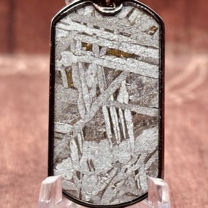 May include: A rectangular pendant with a distinctive, textured pattern. The pendant has a silver-coloured frame and a chain. The surface of the pendant has a complex, interwoven design in shades of grey and silver.