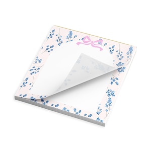 May include: A stack of notepads with a pink floral design. The border features blue floral sprigs and a pink bow at the top. One page is lifted, revealing the blank white paper underneath. The notepad has a gold edge.