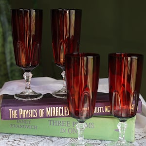 May include: Four tall, deep red glass goblets with vertical fluted detailing on the bowls and clear, faceted stems and bases. The glasses are arranged in two pairs, with two resting on a purple book titled The Physics of Miracles.