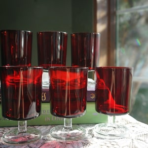 May include: A set of six deep red glass goblets with clear glass stems and circular bases. The glasses feature a smooth, cylindrical bowl design and are arranged in two rows, with three glasses in the front and three glasses in the back.