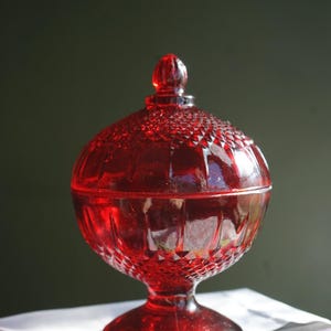 May include: A vibrant red glass candy dish with a lid, featuring a textured diamond pattern on the top and base, a smooth midsection, and a pedestal foot. The lid is topped with a small, rounded finial, creating a classic, symmetrical silhouette.