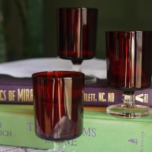Peut inclure: Trois verres à pied en verre rouge profond avec des bols cylindriques, des tiges transparentes et des bases circulaires. Les verres sont disposés en groupe, l'un au premier plan et deux légèrement en retrait, posés sur deux livres reliés empilés.